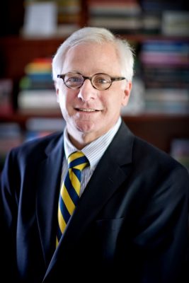 Person with white hair and glasses wearing a suit and striped tie, standing in front of blurred bookshelves.