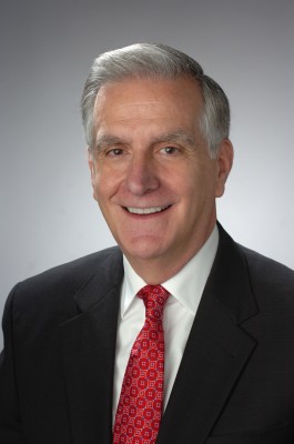 Person with short gray hair wearing a dark suit, white shirt, and red patterned tie against a plain gray background.