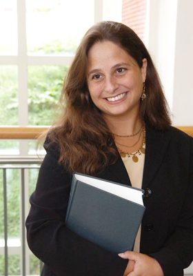 A person with long, wavy hair stands indoors holding a closed book with both hands. They are dressed in a dark suit jacket over a light-colored top. The background includes large windows revealing green foliage outside and metal railings inside.