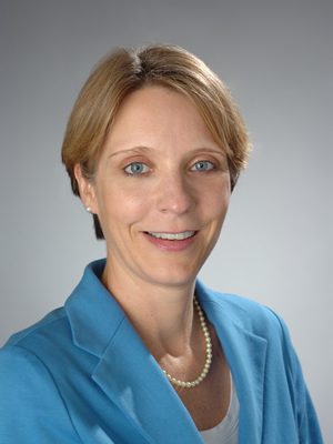 A person with short, light brown hair is wearing a blue blazer and a pearl necklace against a neutral backdrop