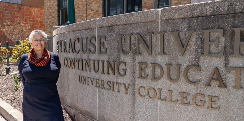 Person smiling beside a stone sign reading “Syracuse University Continuing Education University College,” with a brick building and greenery in the background.