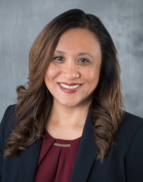 Person with long wavy hair in a dark blazer and maroon top with gold accent, smiling against a light gray background.