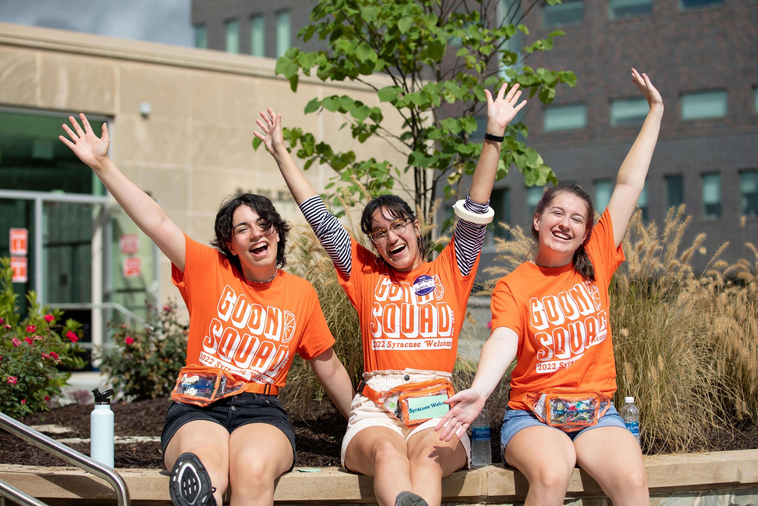Three student volunteers with the Goon Squad sit on a wall with their arms up in the air
