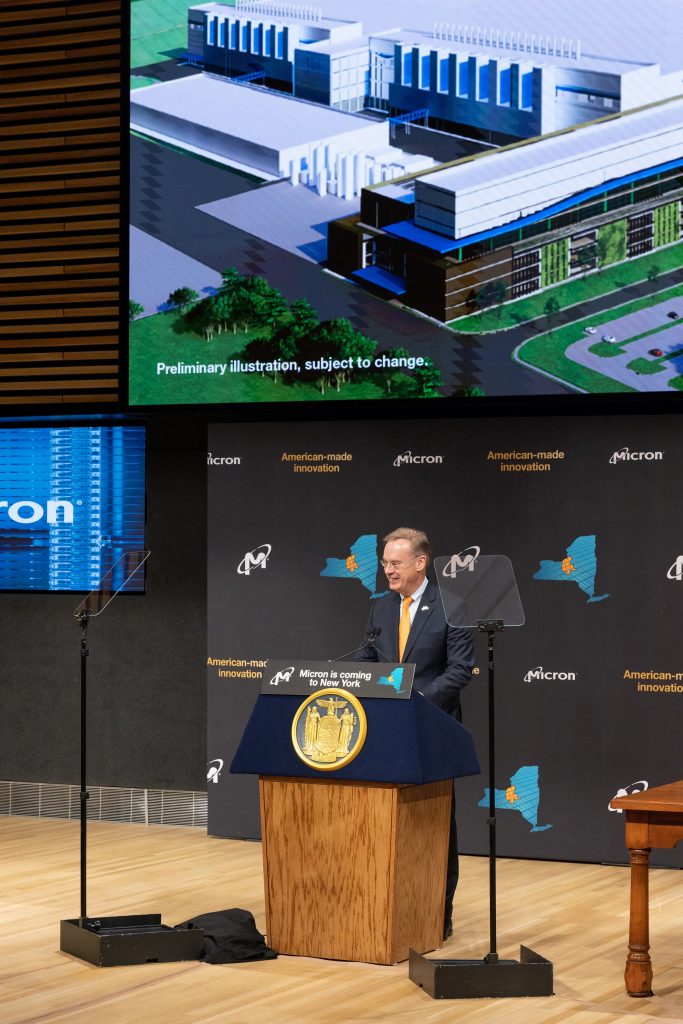 person standing at podium in front of screen showing depiction of manufacturing plant