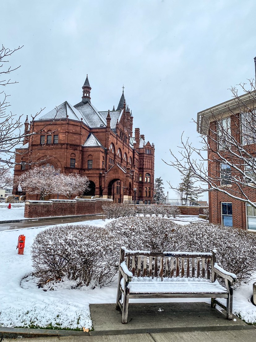 Snow covered campus buildings in the background with a snow covered bench in the foreground. 