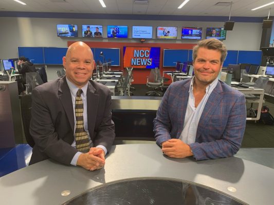 Professors Elliott Lewis and Michael Williams behind a news desk in the Newhouse School