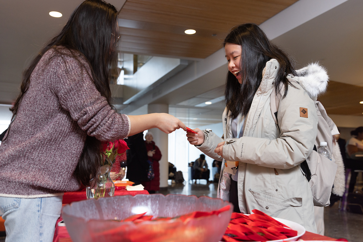 Two individuals standing over a table talking, with one individual handing something to the other. 