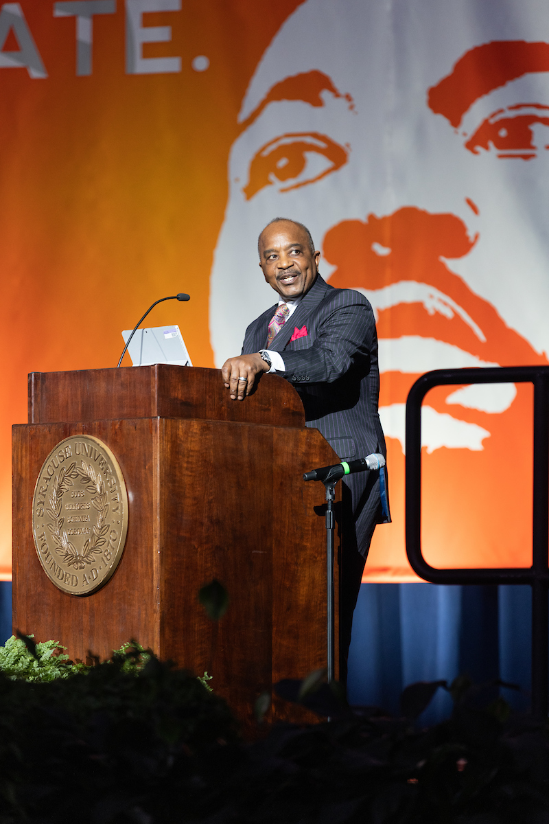 Individual standing at a podium on a stage speaking into microphone. 