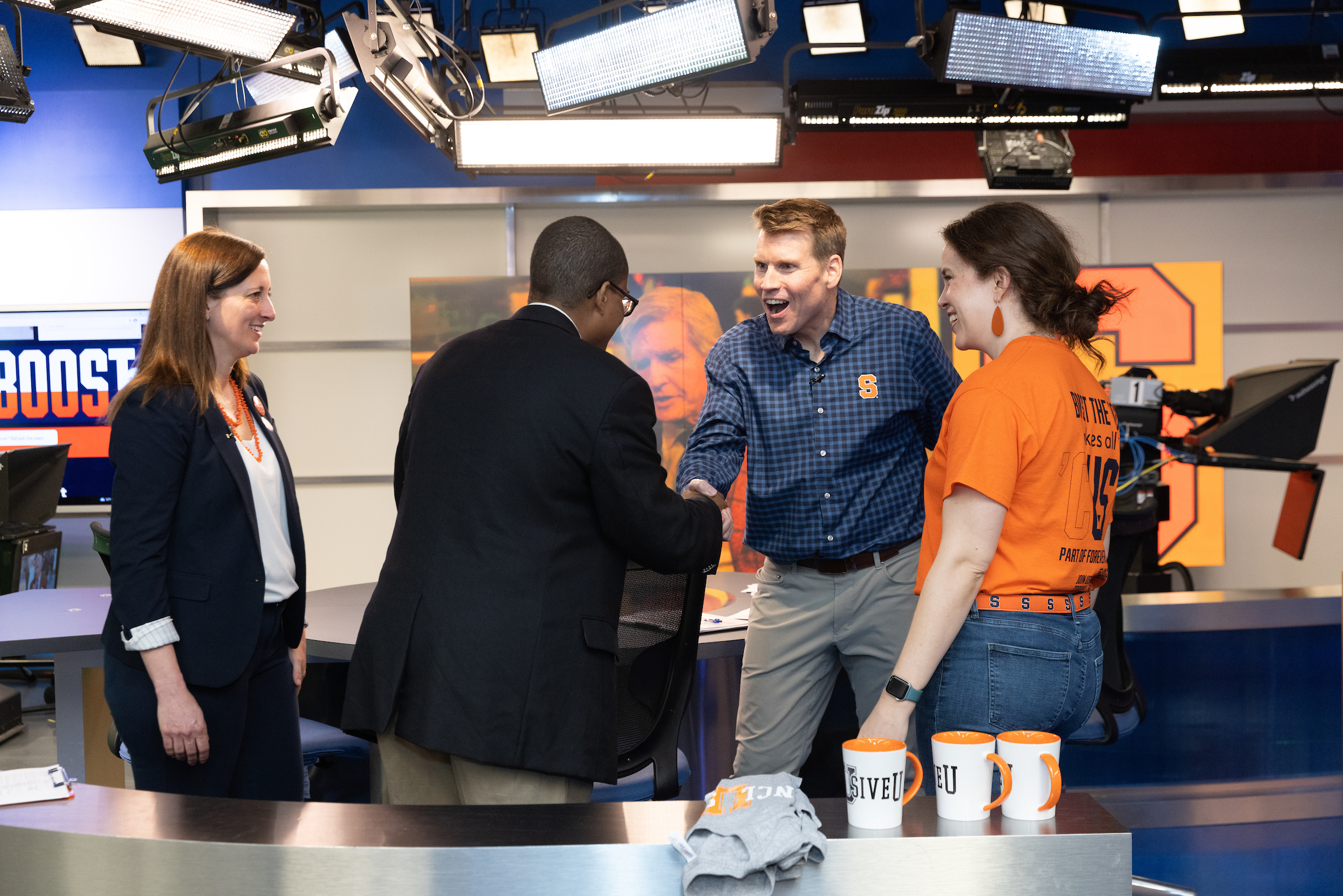 Two men shaking hands, while two individuals stand next to them in a broadcast studio. 