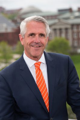 Person in a dark blue suit and orange striped tie standing outdoors with blurred buildings and greenery in the background.