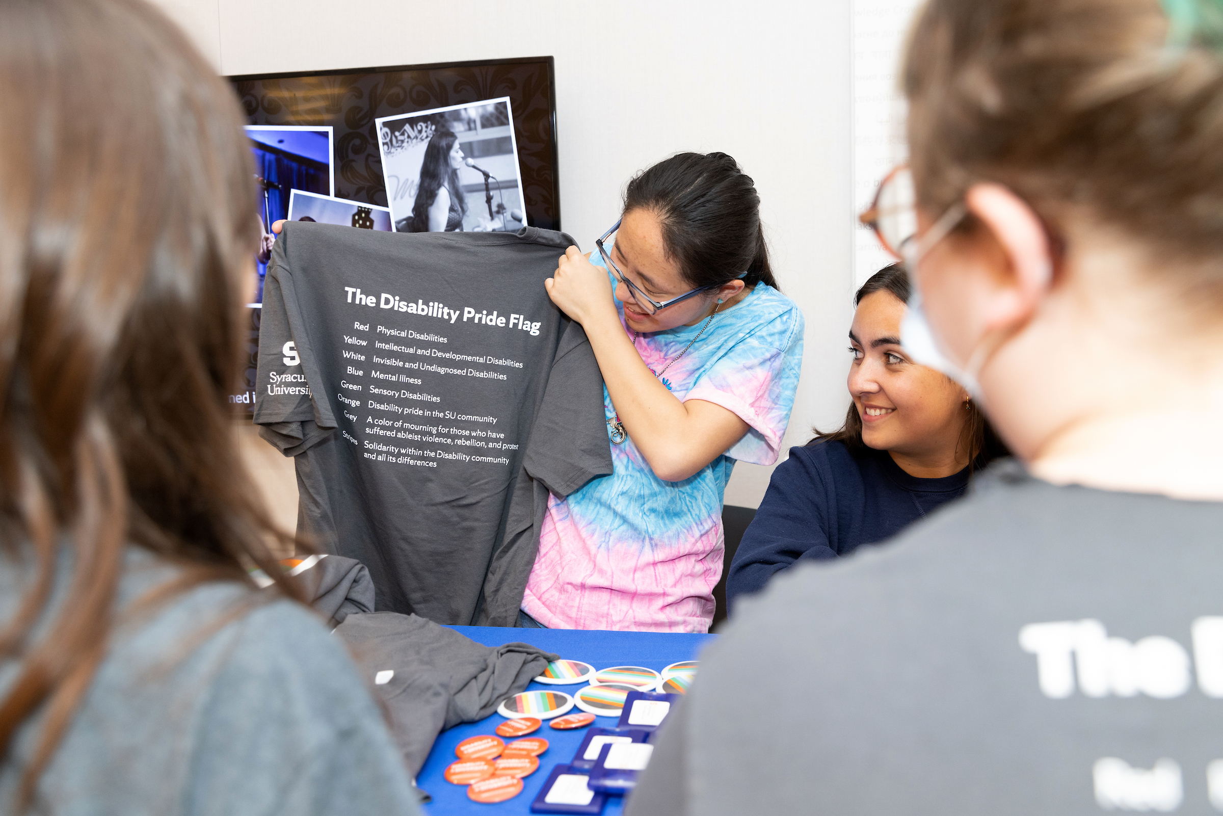 Individual standing holding a grey T-shirt with an explanation of The Disability Pride Flag on the back of it showing it to other students standing nearby.