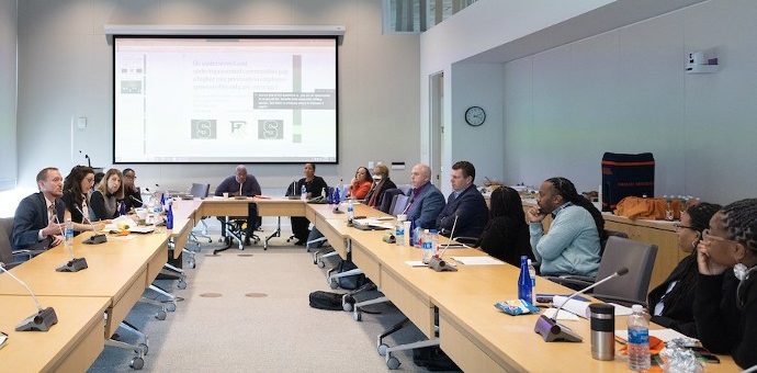 Group of people sitting at tables set up in a rectangle with a presentation on the screen in the front of the room