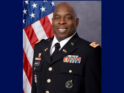 A staff member in military uniform poses for a photo in front of the American flag.