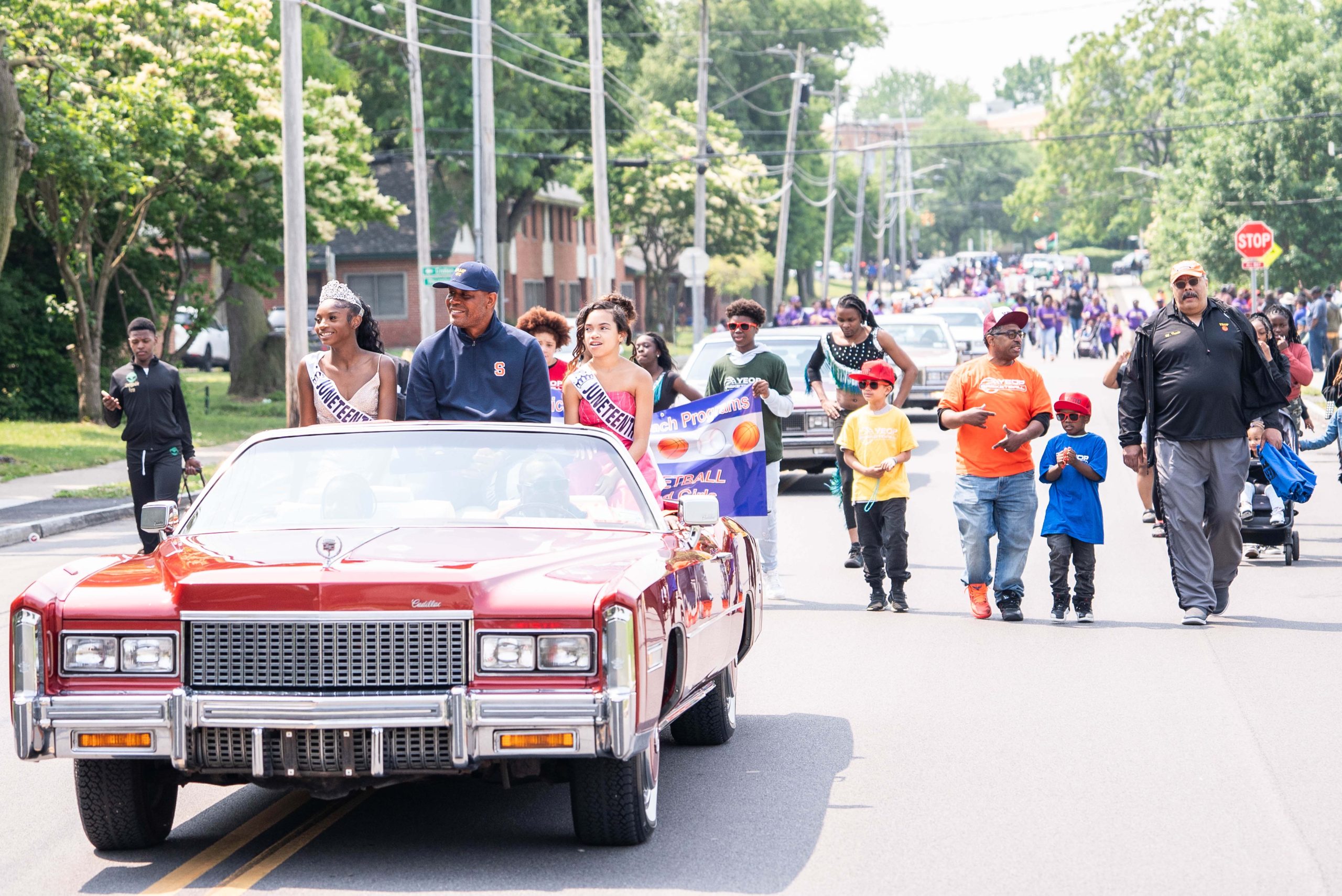 Three individuals sitting in red convertible and others walking behind them in Juneteenth parade.