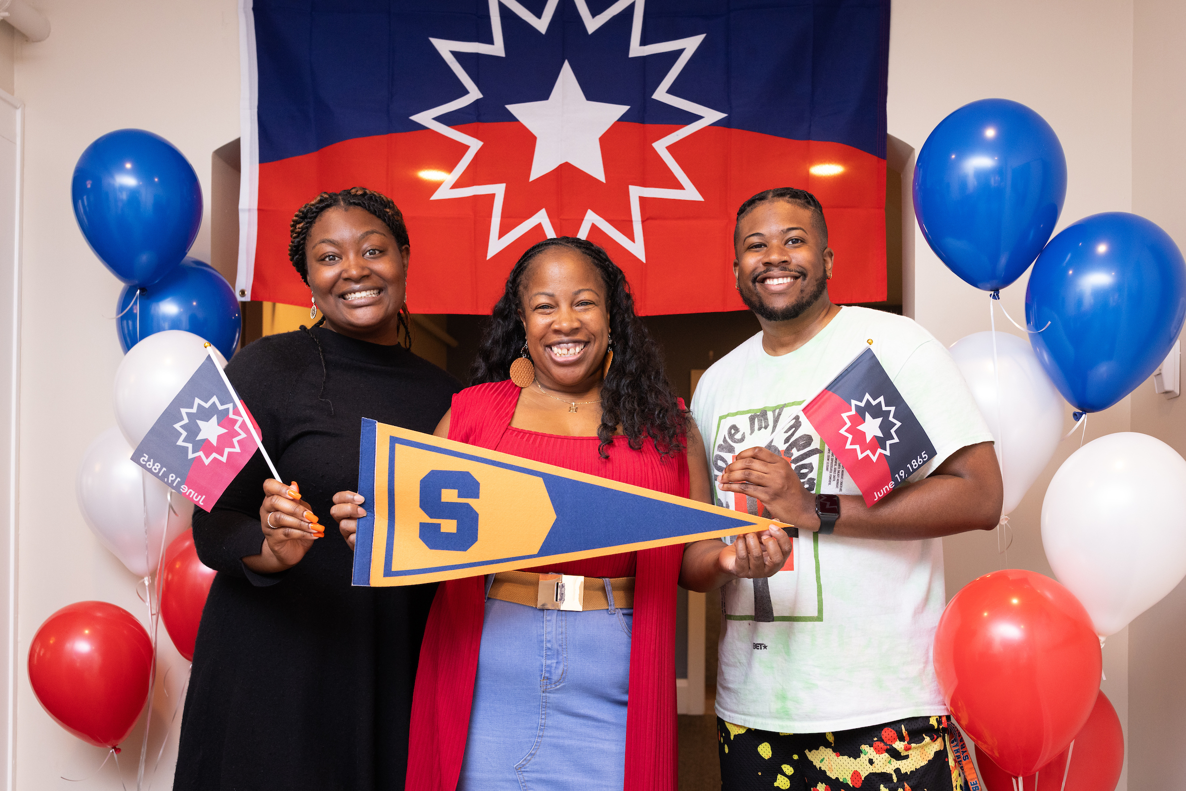 Three individuals standing together in front of flag and red, white and blue balloons during Juneteenth celebration.