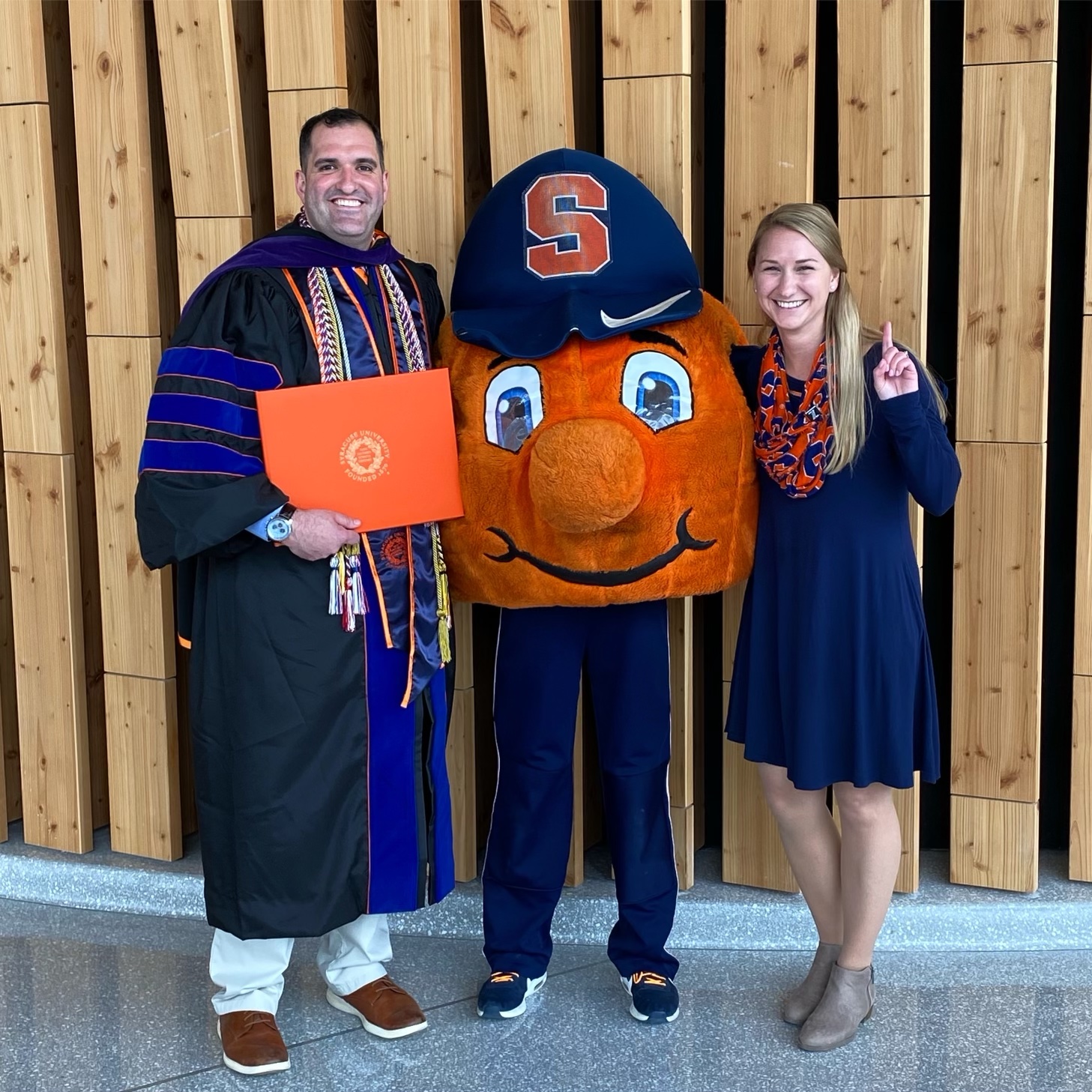 Man in graduation regalia and woman in blue dress standing with Otto 