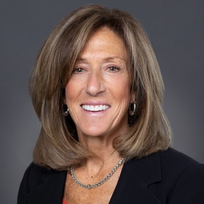 A person with shoulder-length light brown hair, wearing a black blazer and a silver chain necklace, posed against a dark gray background.