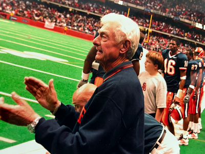 A man stands on the sidelines clapping during a Syracuse University home football game.