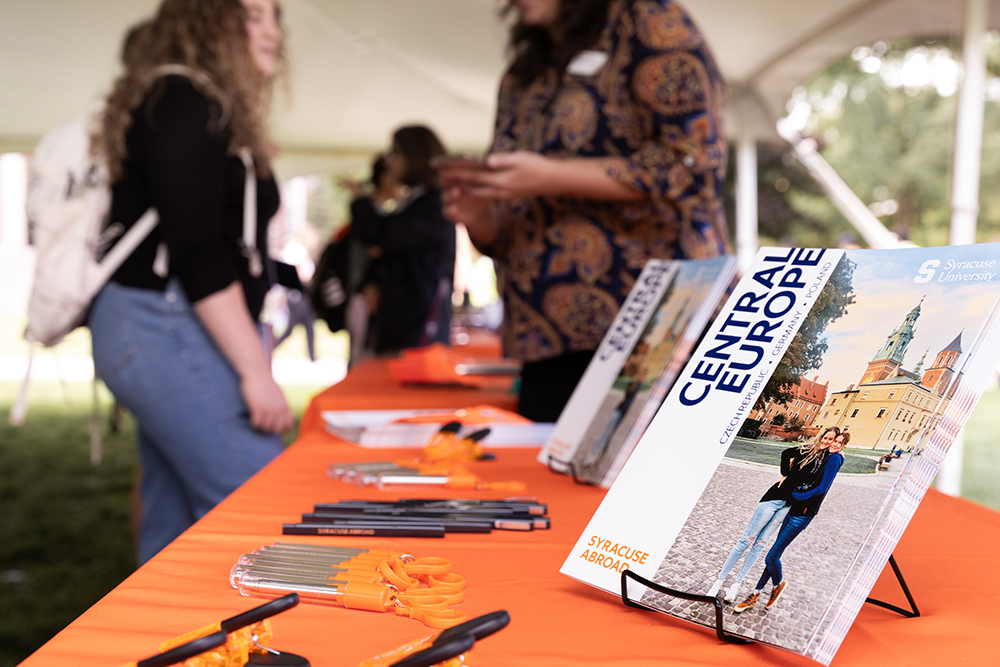 Table under a tent on the Quad with information about Syracuse Abroad with two people talking in the background.