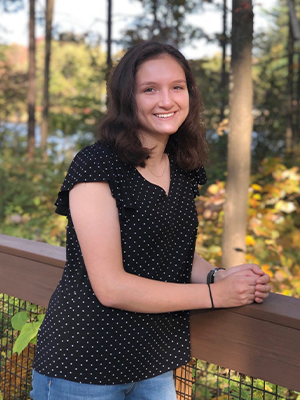 Undergraduate student Alex Snioch poses outdoors against a railing in front of trees and smiles