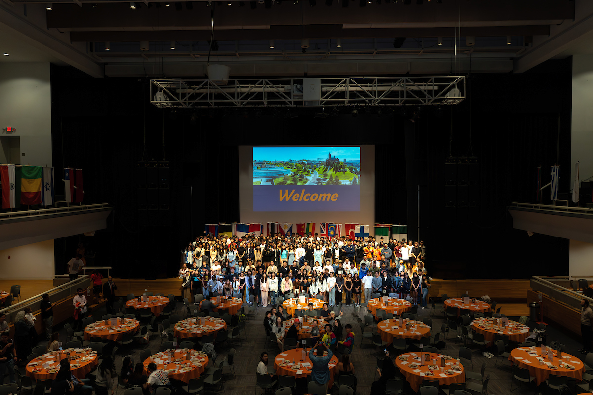 Students pose together in front of a screen that says Welcome and flags from numerous countries at the international student welcome dinner