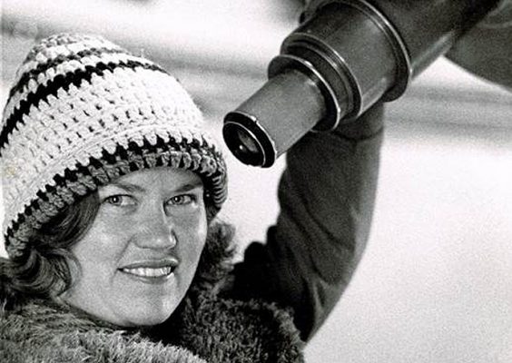 A woman wearing a winter hat poses for a photo while standing next to a telescope.