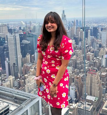 graduate student Meeti Shah poses in front of a window amidst the backdrop of the bustling buildings of New York City