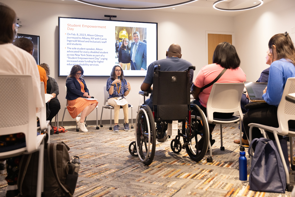 Two individuals sitting in the front of a room in front of a screen with a PowerPoint presentation on it, presenting to an audience.
