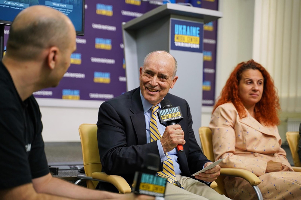 Three people sitting on a stage. One with a microphone speaking.