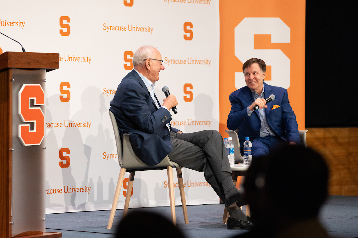 Two men sitting on a stage with microphones speaking.