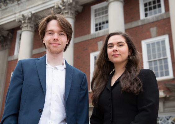 A man wearing a blue sport jacket and white dress shirt stands next to a woman wearing a black blazer outside of the Maxwell School of Citizenship and Public Affairs on the Syracuse University campus.