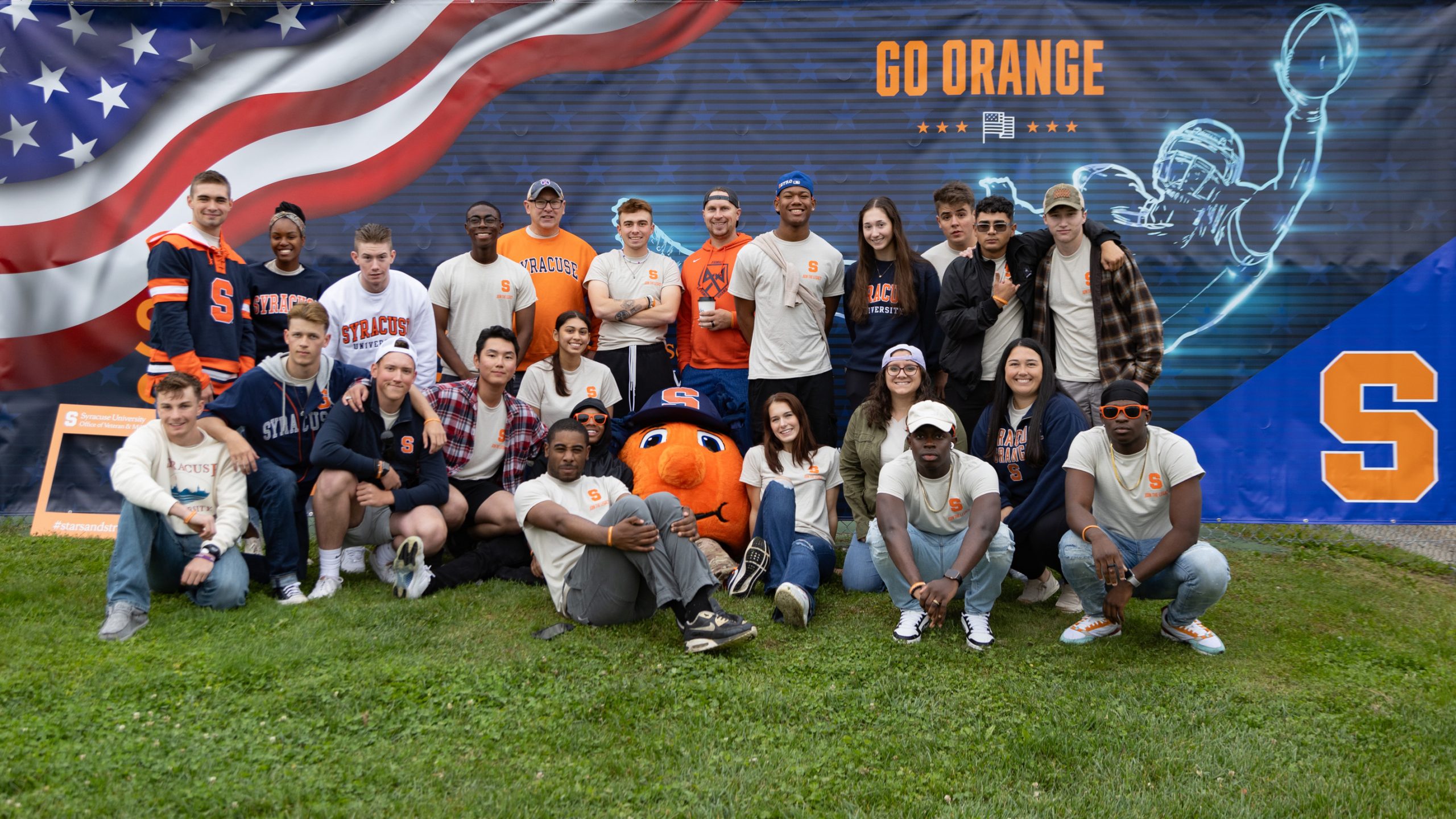 Group of students standing together with Otto in front of the Stars & Stripes banner. 
