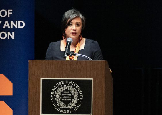 A woman stands at a podium with the Syracuse University shield and motto during the DEIA Symposium.
