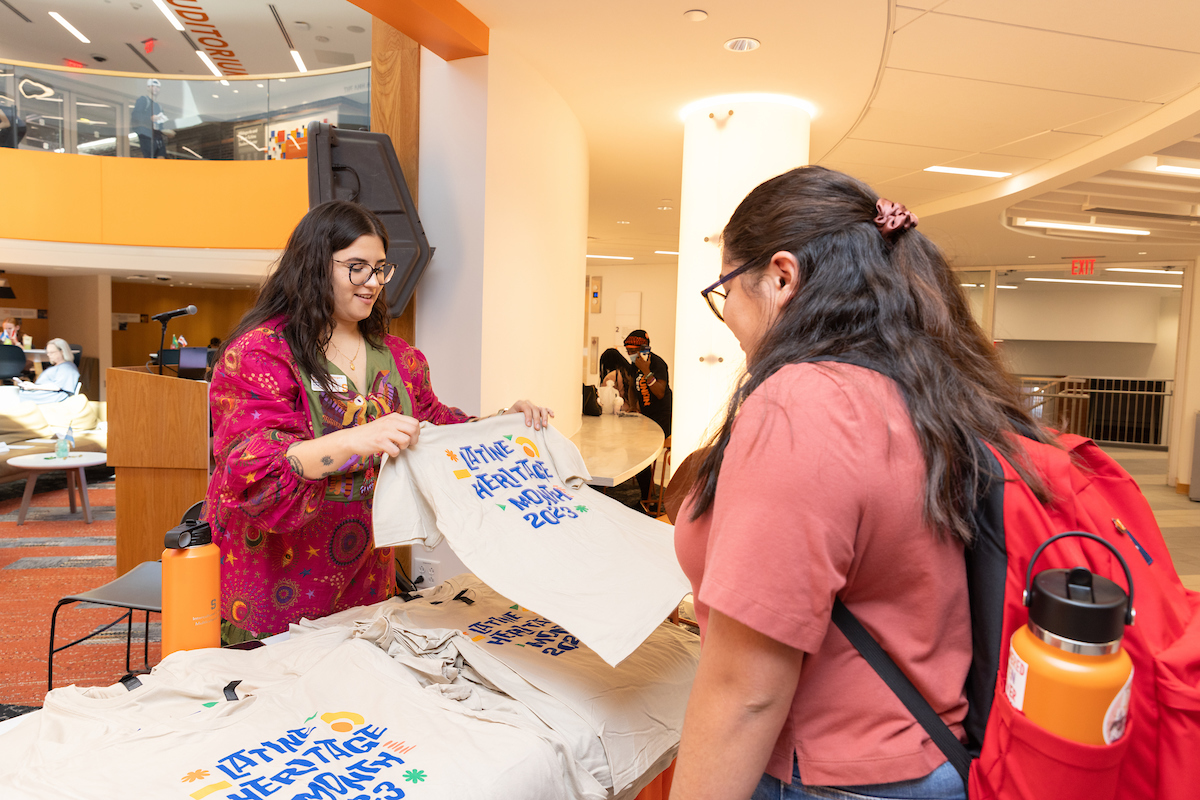Individual standing at a table filled with t-shirts that say Latine Heritage Month 2023 with a student standing looking at the shirts. 