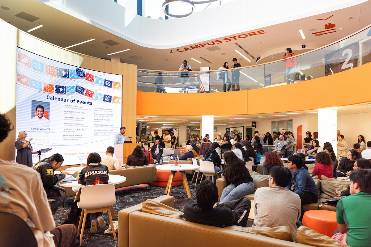Large group of students in the Schine Student Center watching someone speaking in front of the large screen. 