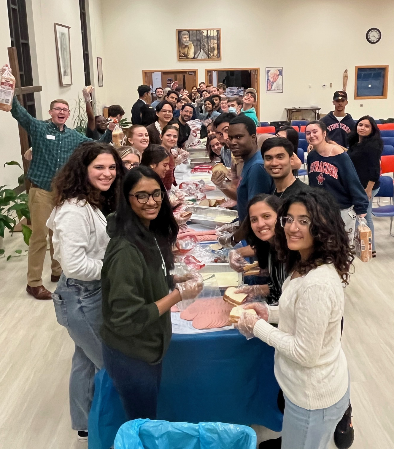 Large group of people gathered on either side of a table making sandwiches.