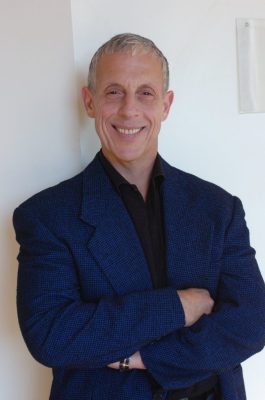 Person with short light hair in a dark blue blazer and black shirt, smiling with arms crossed against a white wall.