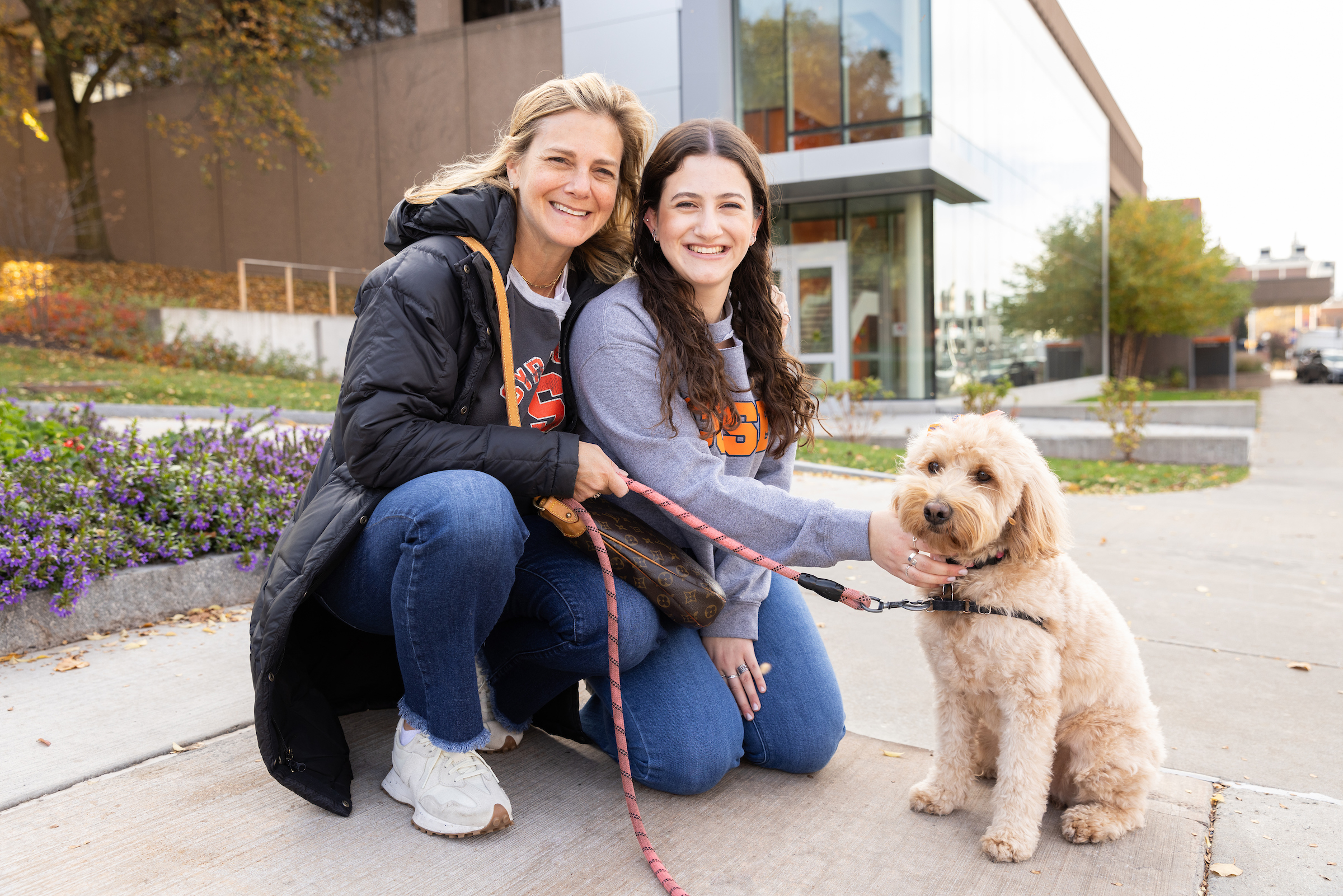 Two individuals in Syracuse gear pose with a dog on campus