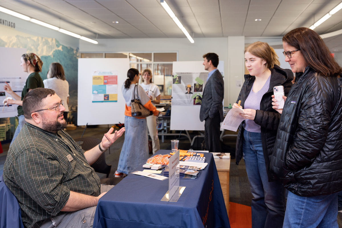 Two individuals speak to a person seated at a table during the Family Weekend Honors Residential Reception in Sadler Hall