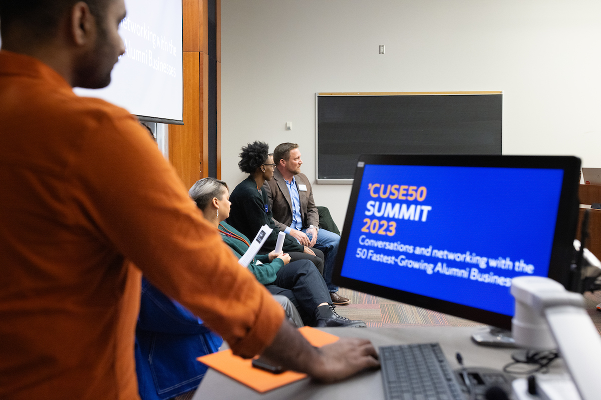 Several individuals sitting at the front of a classroom for a panel discussion, with someone standing at the adjacent computer station. 