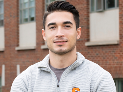 A man poses for a photo while standing outside wearing a grey Syracuse zip up.