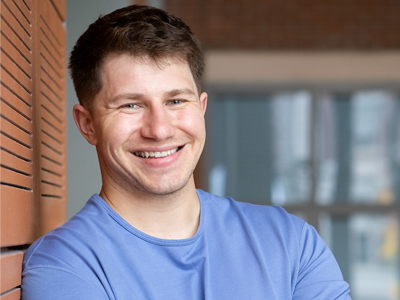 A man smiles for a headshot while leaning against a wall.