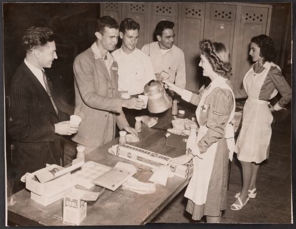 Black and white photo of two women from the Red Cross serving people standing on the other side of a table.