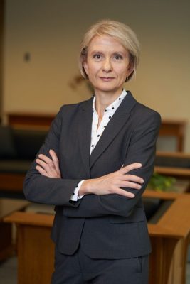Person in a dark gray suit and polka dot shirt standing with arms crossed in an office setting.