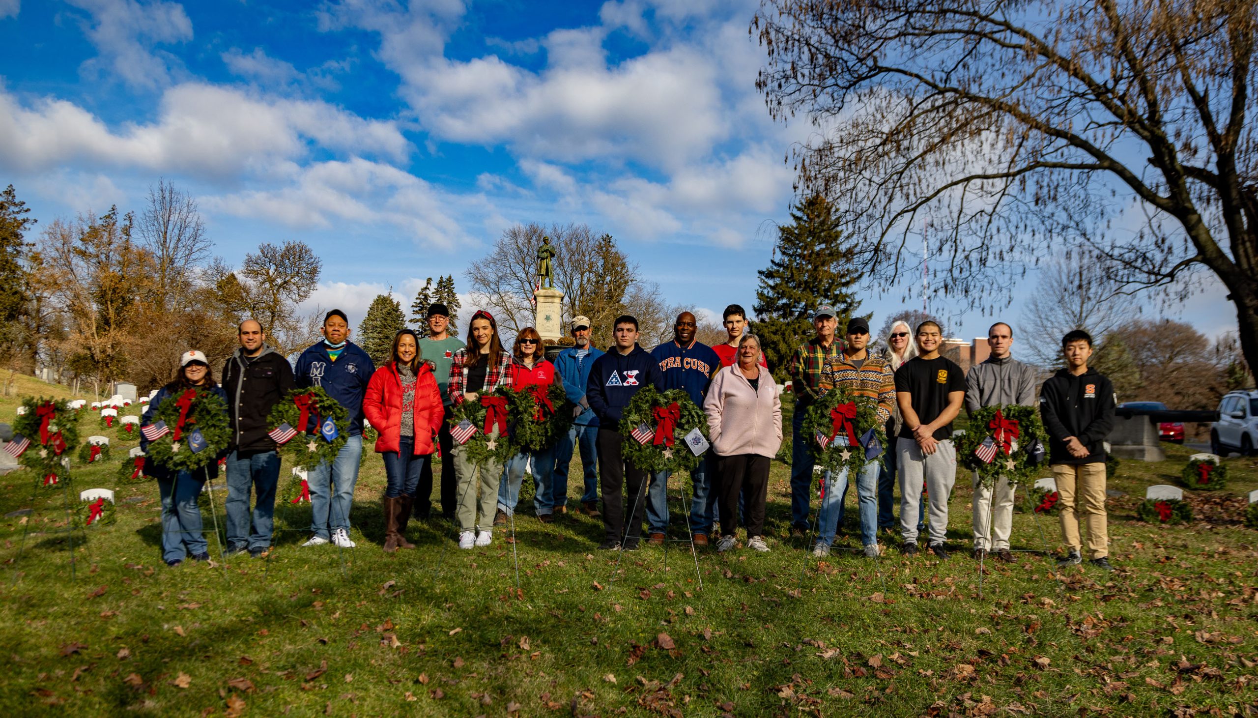 Group of people standing in a cemetery with christmas wreaths in front of them on a sunny day.