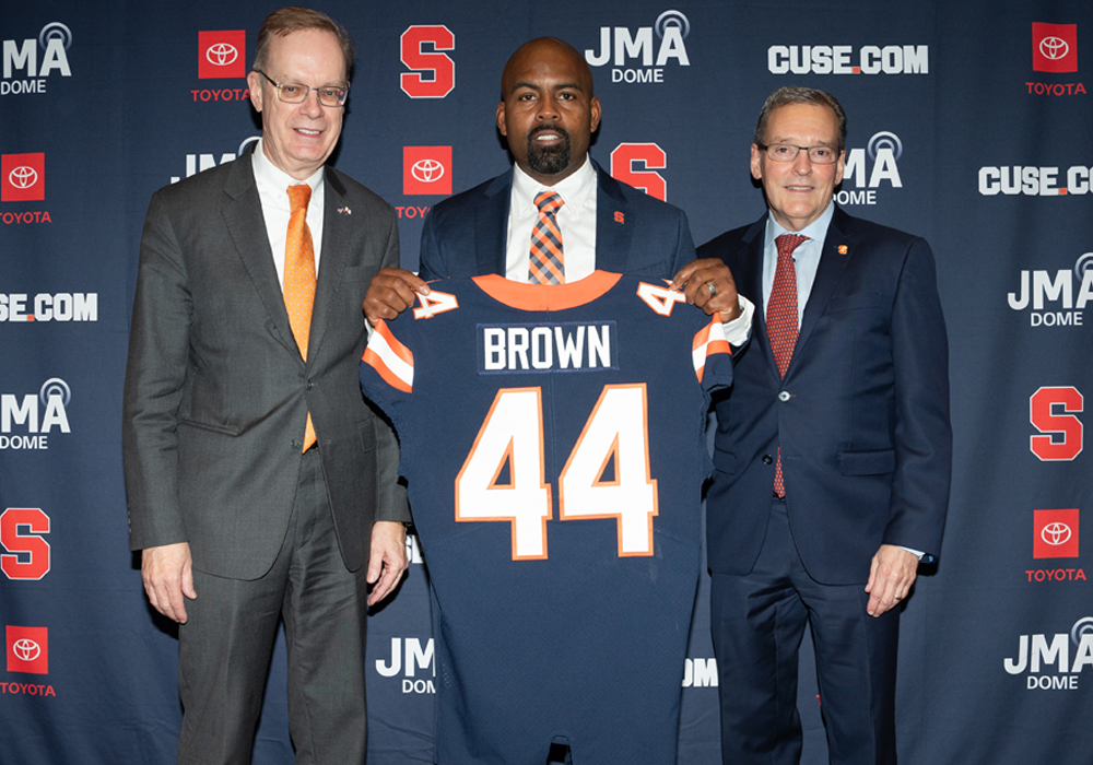 Three men pose for a photo while holding up a blue Syracuse University number 44 football jersey