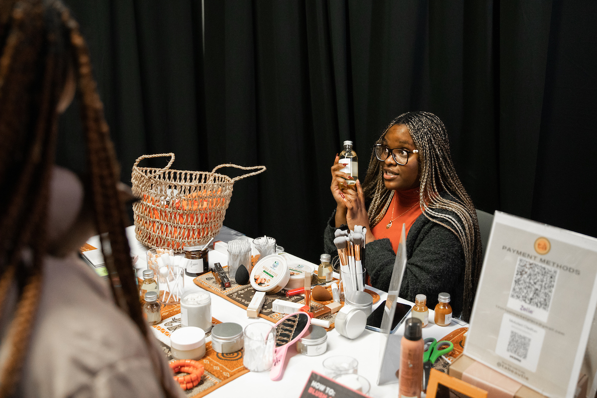 Individual sitting at a table with beauty products talking to someone on the other side of the table. 