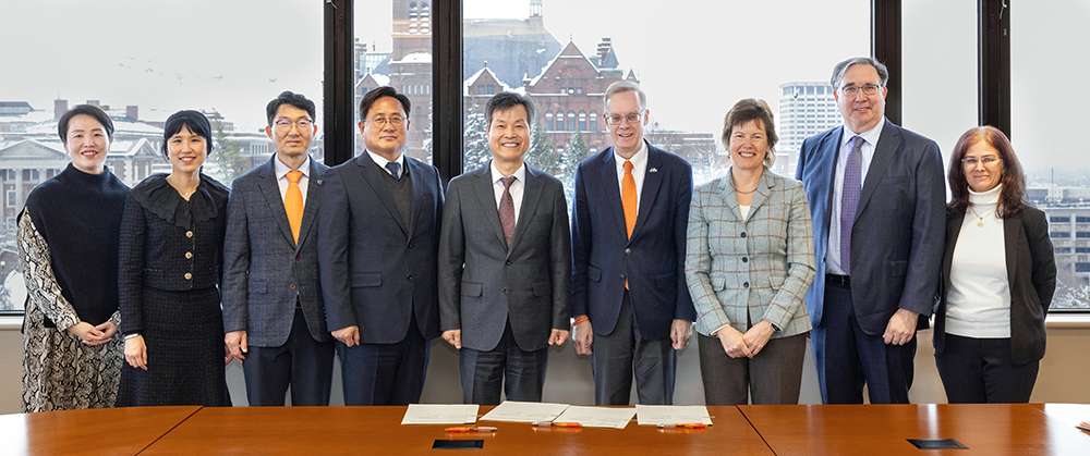 9 individuals representing the University of Seoul and Syracuse University pose together behind tables and in front of windows overlooking a snowy campus