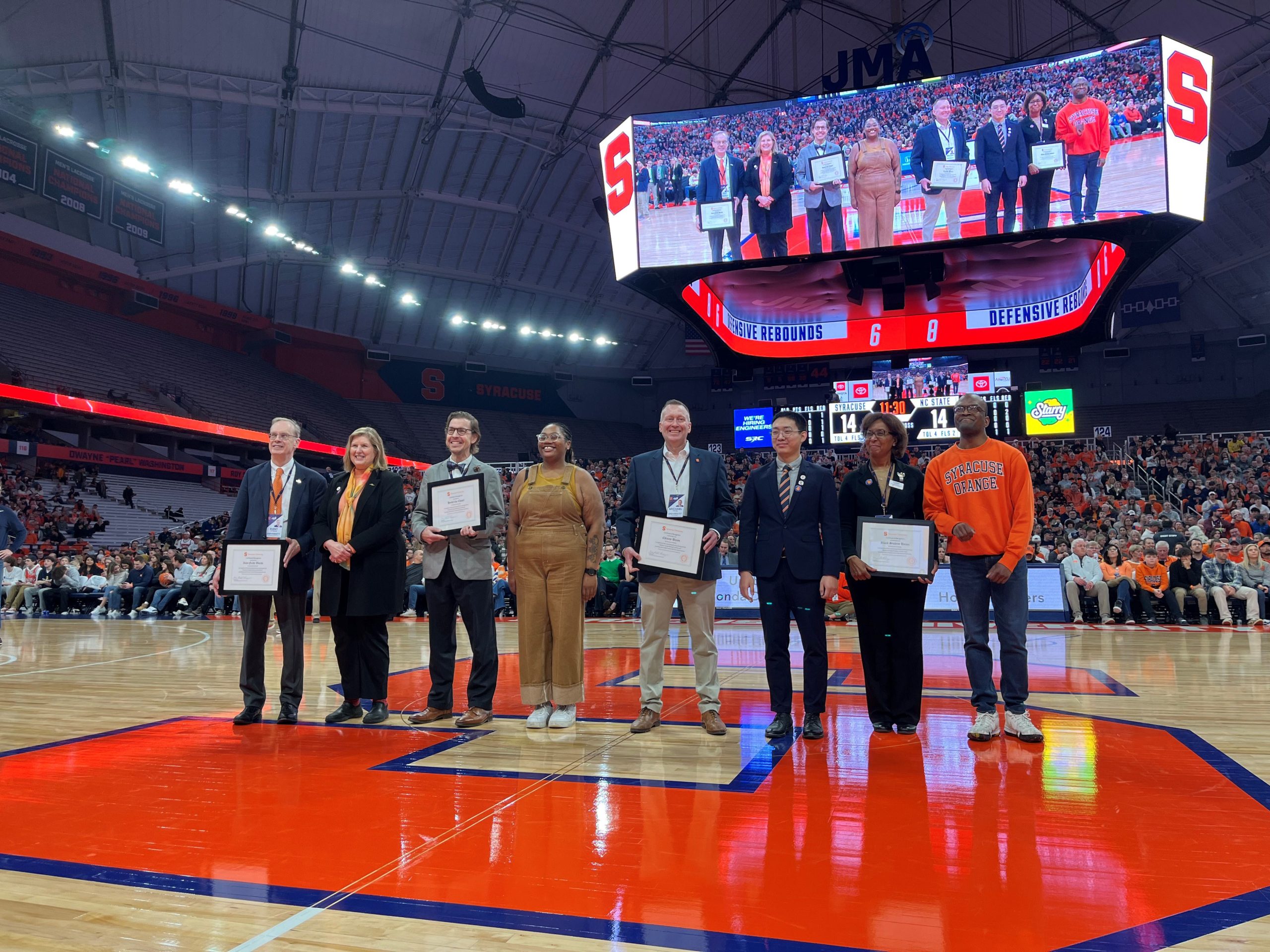 Group of eight people standing on the court at the JMA Wireless Dome during a recognition ceremony.