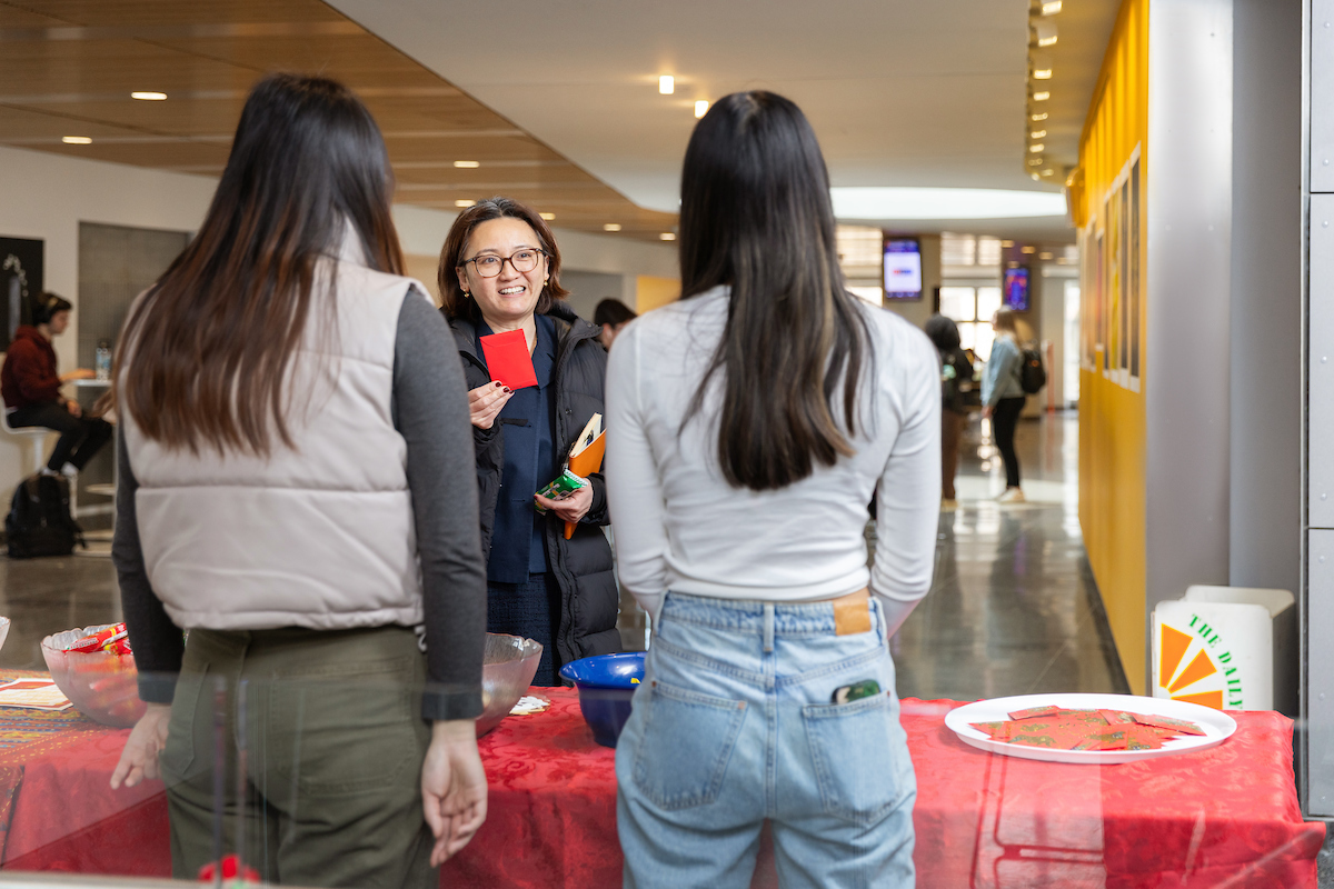 Two people standing with the backs to a camera talking to a another person facing them on the other side of a table.